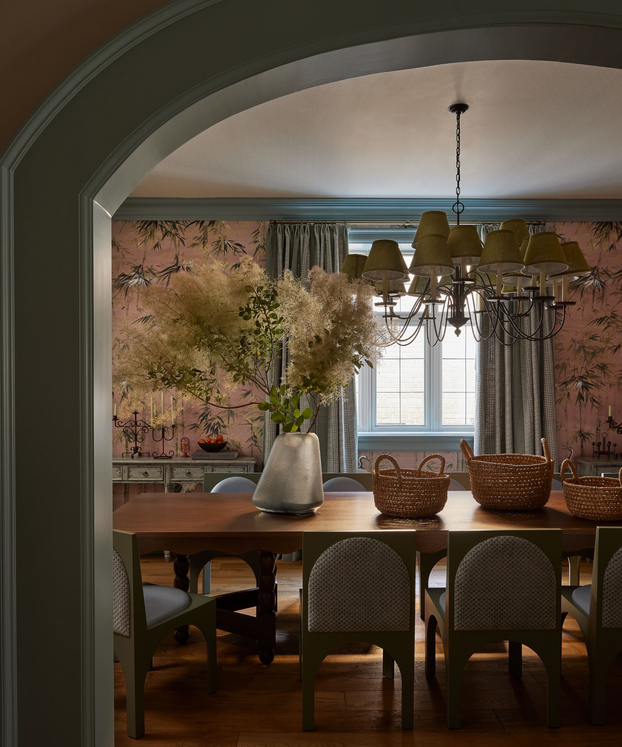 Arch doorway looking through to a dining room, blue painted trim, pink patterned wallpaper on walls, large wooden dining table with upholstered chairs, vase of flowers and baskets on table