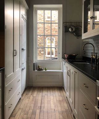 Galley kitchen with cream cabinets and narrow wooden floor boards in front of a large sash window