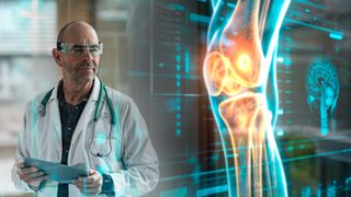 A bald man with clear glasses wearing a white lab coat and stethoscope looks at a holographic blue and orange image of a leg and leg bone.