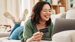 A happy woman lying on her stomach on a sofa using her payment card to buy something on her laptop. We cannot see the screen