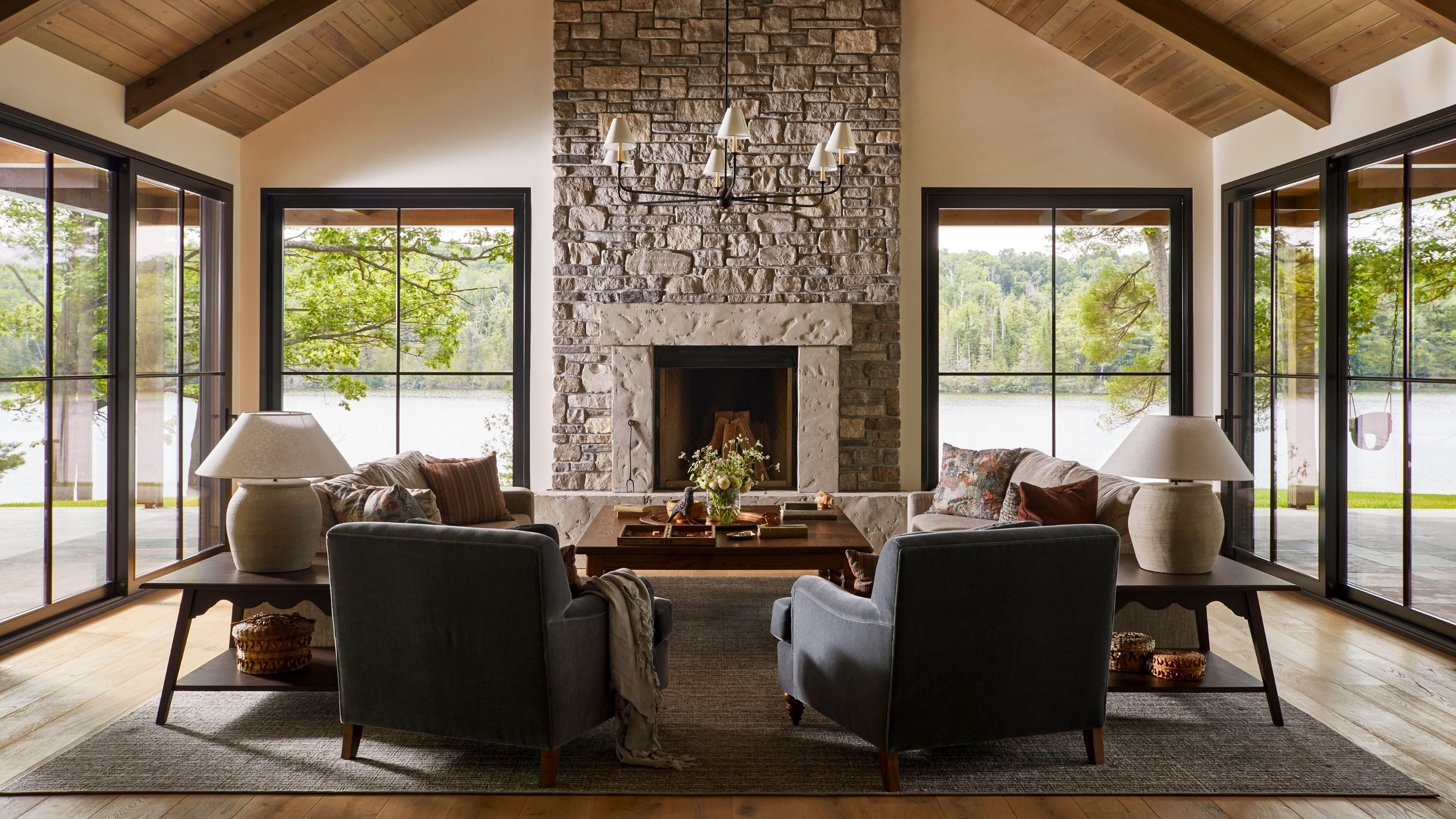 A spacious living room with vaulted wood ceilings, floor-to-ceiling windows, and a massive floor-to-ceiling stone fireplace as the centerpiece.