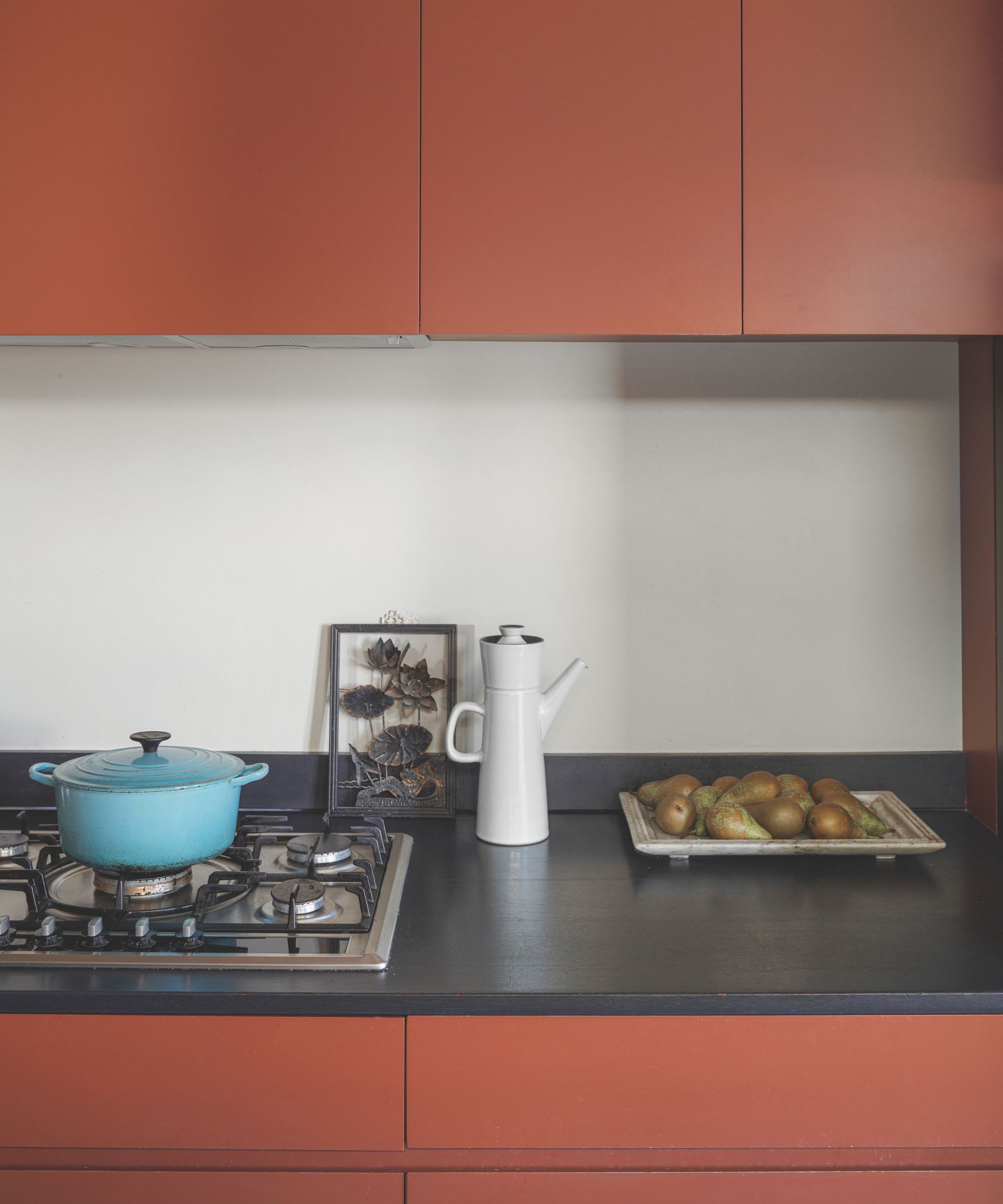 A orange kitchen with orange cabinets, black countertop, white backsplash, stove, blue pan, white kettle and fruit bowl filled with pears.