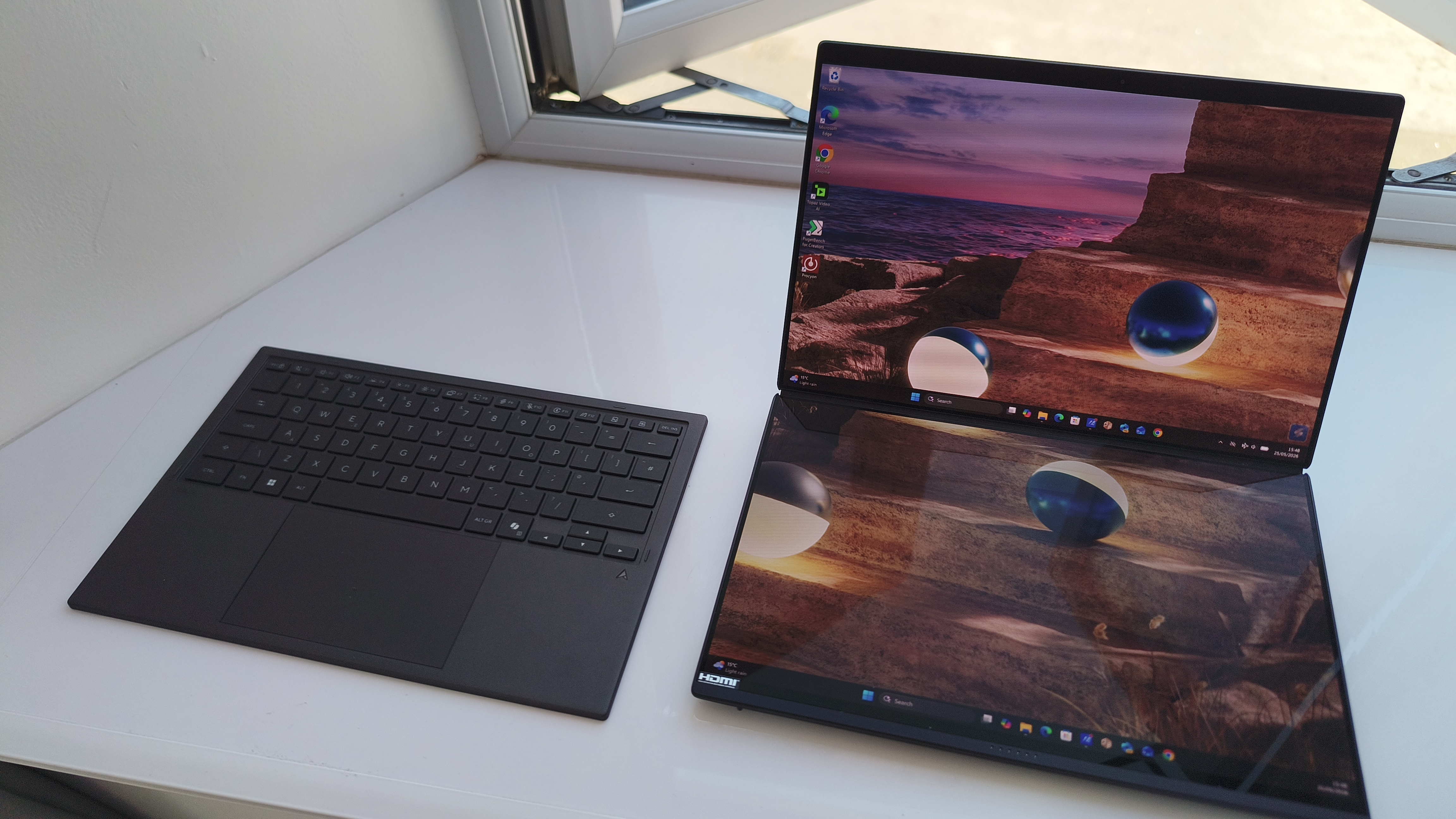 The ASUS Zenbook Duo propped open on a white counter by a window to reveal both of its 14-inch landscape screens, with its slim, dark grey wireless keyboard detached and resting to the left.