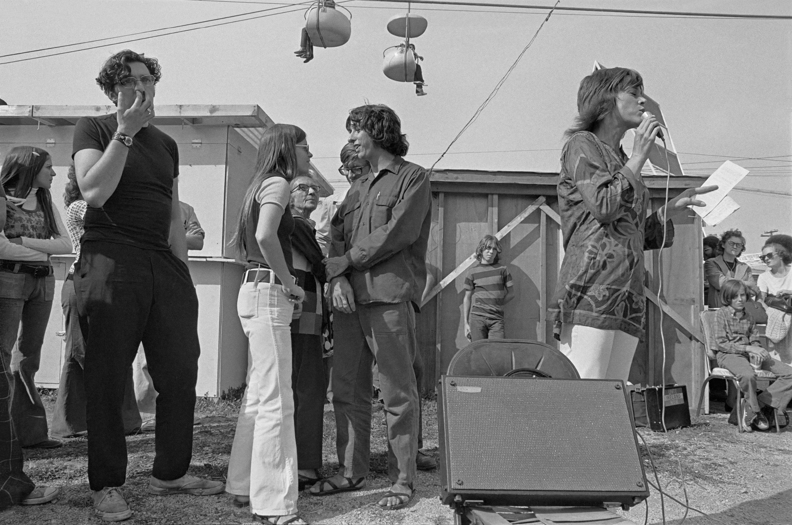 Jane Fonda speaks into a microphone at an outdoor anti-war rally as people stand talking nearby and fairground ride seats hang from cables overhead.