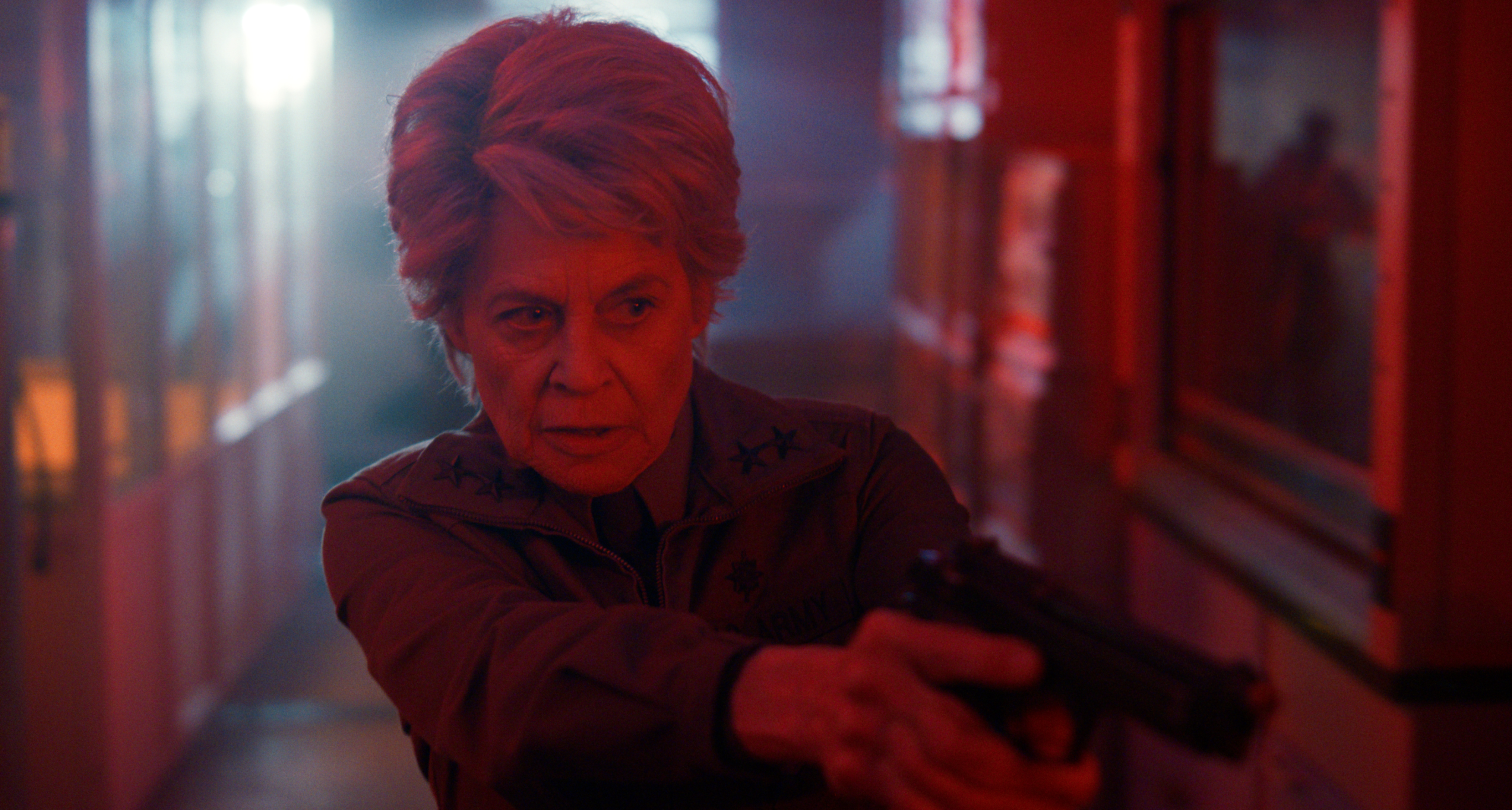 linda hamilton as dr. kay pointing a gun walking down a dimly red lit hallway in stranger things season 5