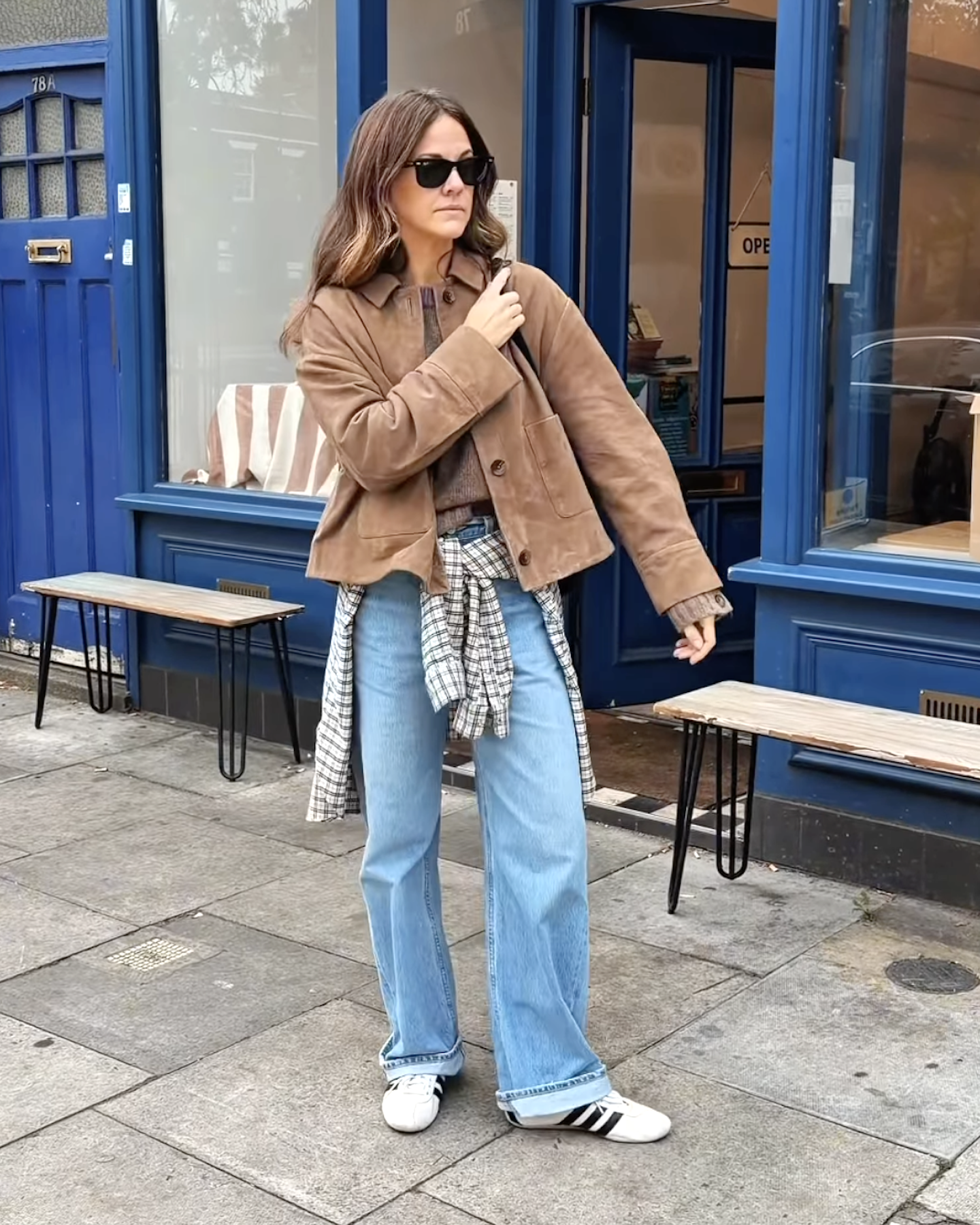 British style influencer Kim Turkington poses on a London street wearing a tan suede jacket, a plaid shirt tied around her waist, cuffed jeans, and Adidas Tokyo sneakers.