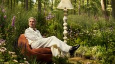 Designer Tan France poses sitting atop of a vintage-inspired accent chair in a whimsical forest setting alongside a sculptural monochrome floor lamp