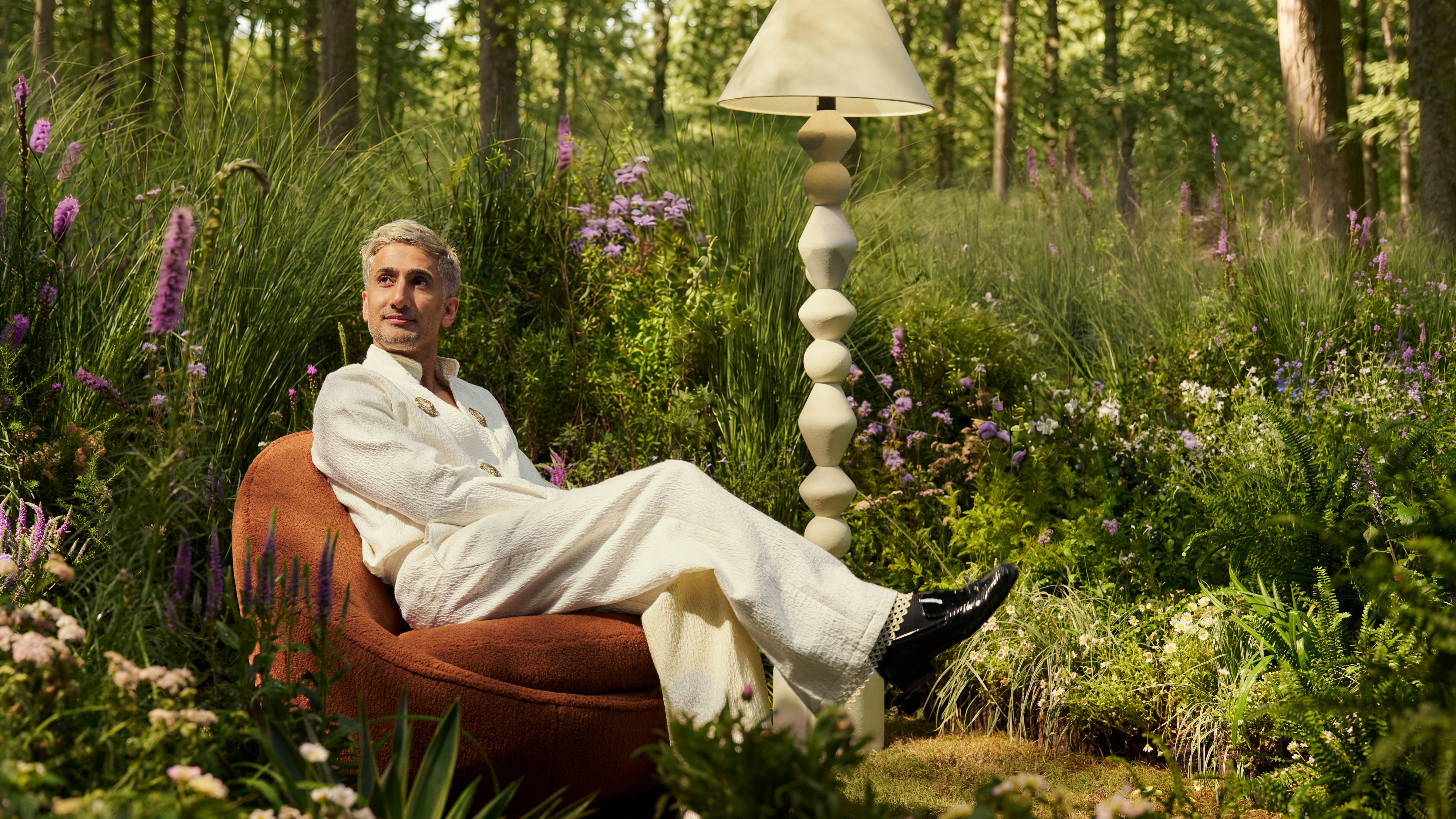 Designer Tan France poses sitting atop of a vintage-inspired accent chair in a whimsical forest setting alongside a sculptural monochrome floor lamp