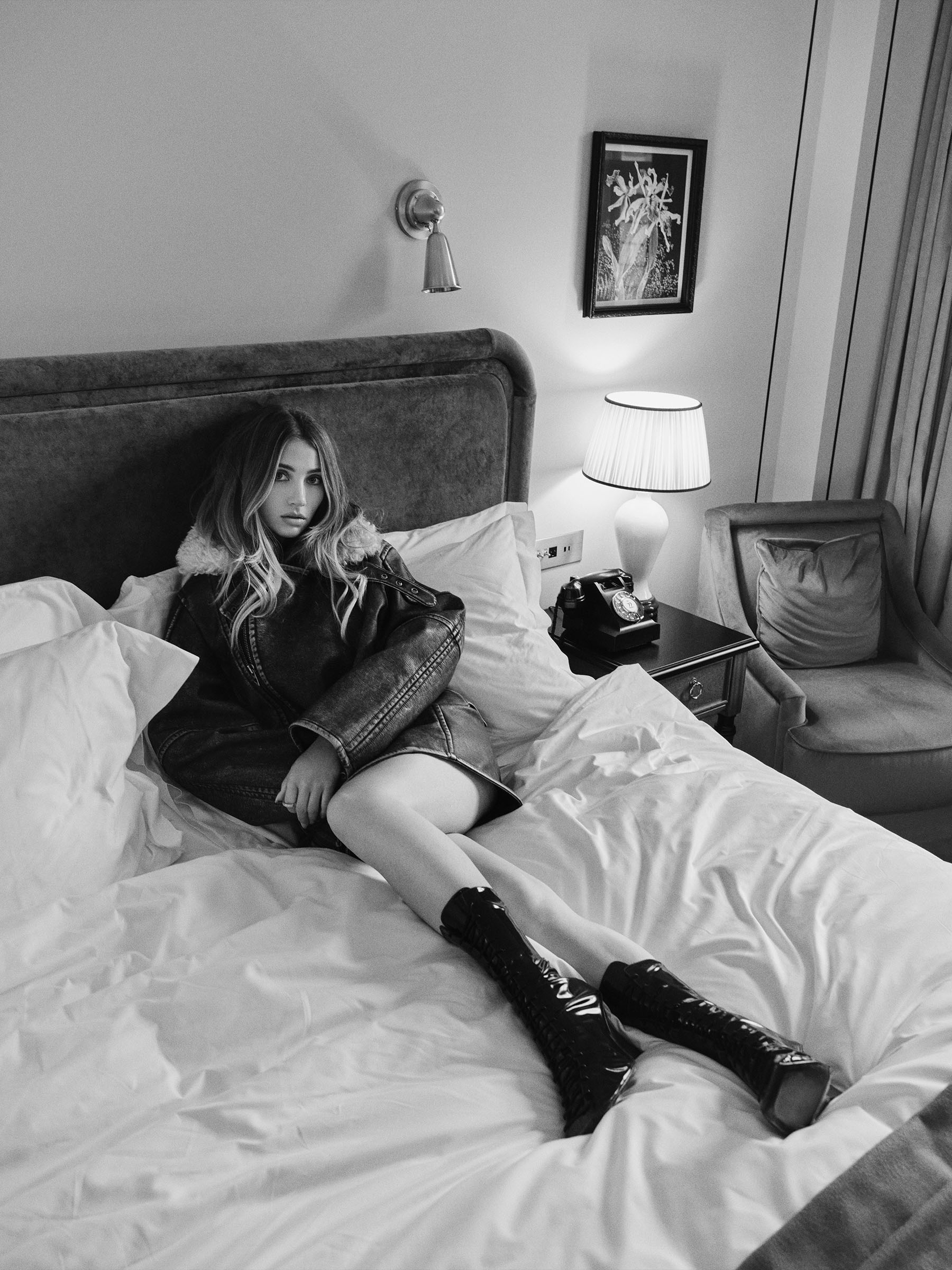 Emily Rudd photographed by Christian H&amp;ouml;gstedt. She is lying in bed in a hotel room wearing a leather jacket with fur collar and black patent leather boots.