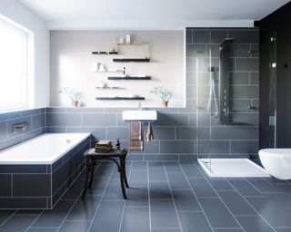 Beautiful dark blue bathroom with white sink, bath and toilet and blue tiled floor and walls, a vintage wooden side table with books on top, with light shining in from bright window to left side of picture
