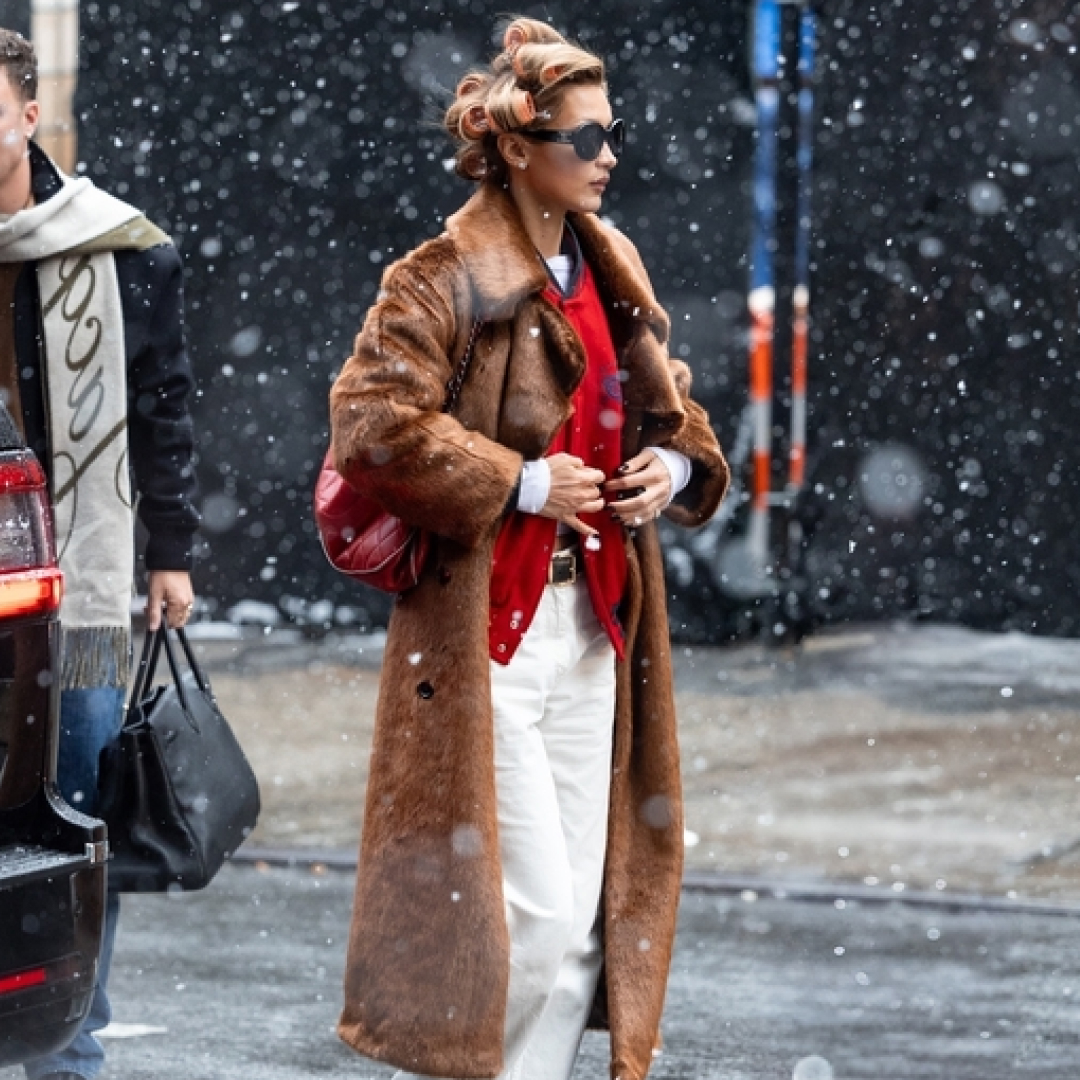 Bella Hadid wearing a long fur coat with a red sweater, white jeans, and black sneakers