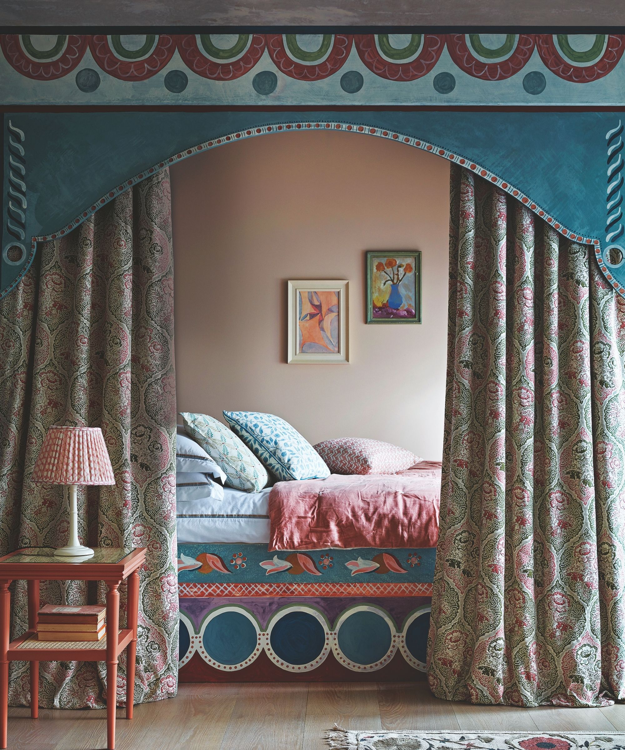 A colorful, patterned canopy bed in a peach bedroom with patterned curtains, pink bedding, patterned cushions, orange bedside table with a white and red lamp, and light wood flooring.
