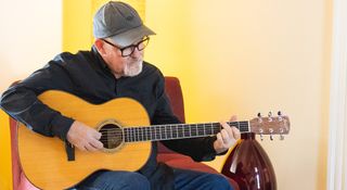 Session pro Tim Pierce is photographed at home, wearing a ballcap and fretting a chord on his guitar.