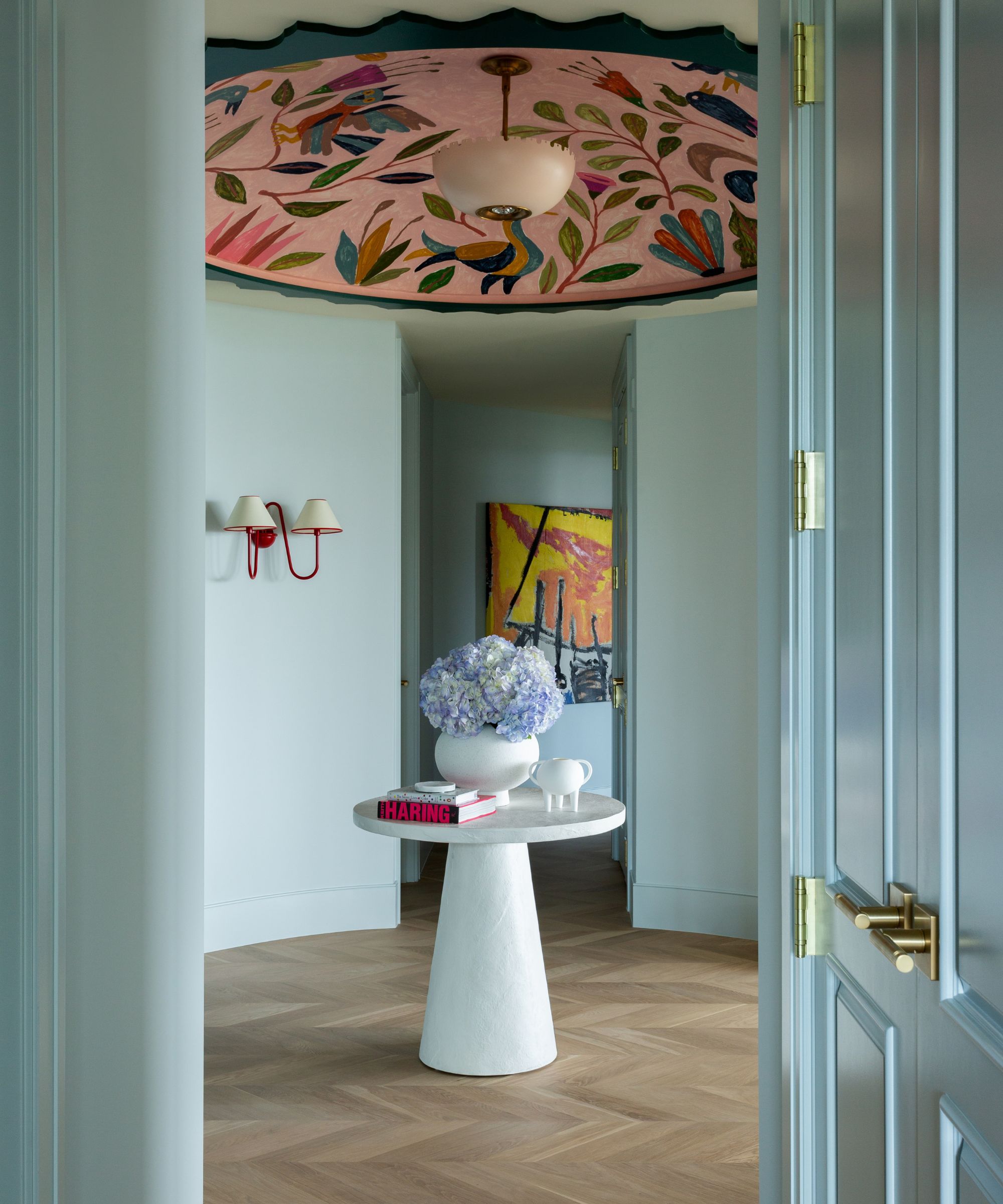 Pale blue entryway with dark teal double doors, two red sconces on walls, a large pink floral ceiling mural, a round, small white table in the centre with a vase and books, and parquet wooden floors