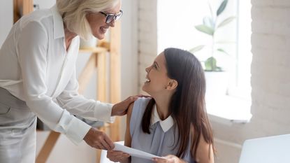 The boss hands an envelope with her bonus in it to a woman sitting at her desk in an office.