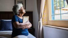 An older woman holds a framed photo to her chest while she looks out her bedroom window.