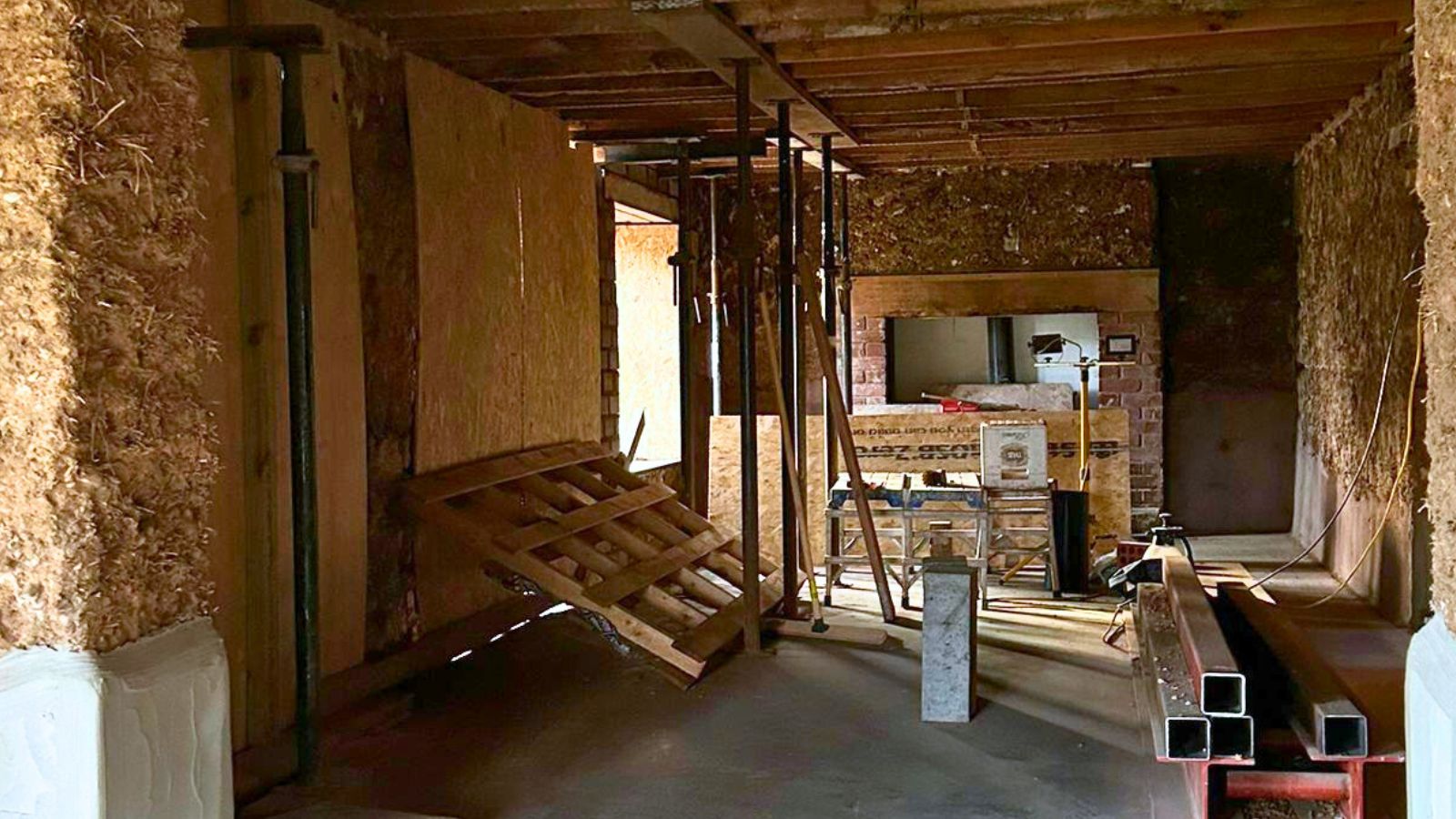 A large room mid-renovation with exposed timber ceilings, straw-filled cob walls and multiple acrow props supporting new openings