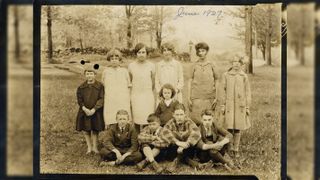 A faded sepia photograph of nine children and a young teacher posing on a grassy lawn in front of a stone wall and trees, with four boys sitting cross-legged at the front, the girls standing behind