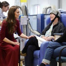 Kate Middleton sitting in a chair wearing a red sweater and skirt putting her hand on the knee of a cancer patient wearing a cold cap getting chemo treatment