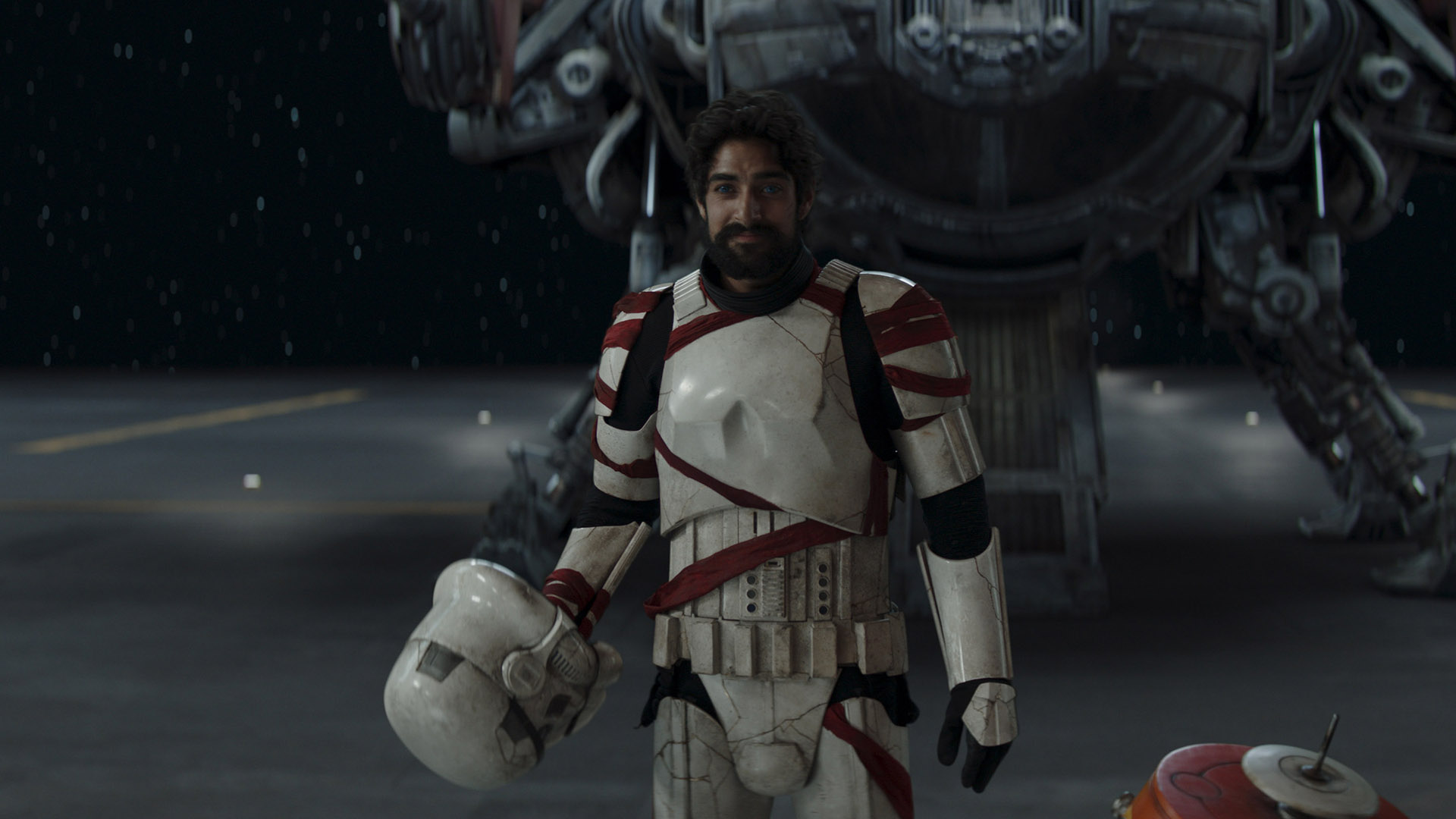 Still from season 1 of the Star Wars TV show Ahsoka. Ezra returns (human male with short dark hair and a full dark beard). He is wearing white stormtrooper's armor, wrapped here and there with red tape. He has just removed his helmet.