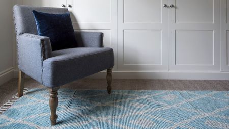 A blue upholstered chair with brown legs and a dark velvet cushion in front of some white cupboards with wooden flooring and a blue patterned rug.
