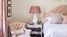 Pink bedroom with a patterned pink and white headboard, cushions, wallpaper, and curtains. The bedding is white, and there's a white nightstand, pink and white lamp, and pink upholstered chair.