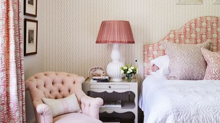 Pink bedroom with a patterned pink and white headboard, cushions, wallpaper, and curtains. The bedding is white, and there's a white nightstand, pink and white lamp, and pink upholstered chair.