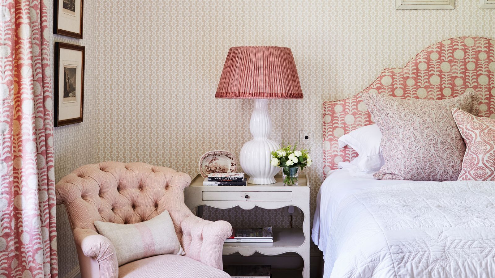 Pink bedroom with a patterned pink and white headboard, cushions, wallpaper, and curtains. The bedding is white, and there's a white nightstand, pink and white lamp, and pink upholstered chair.