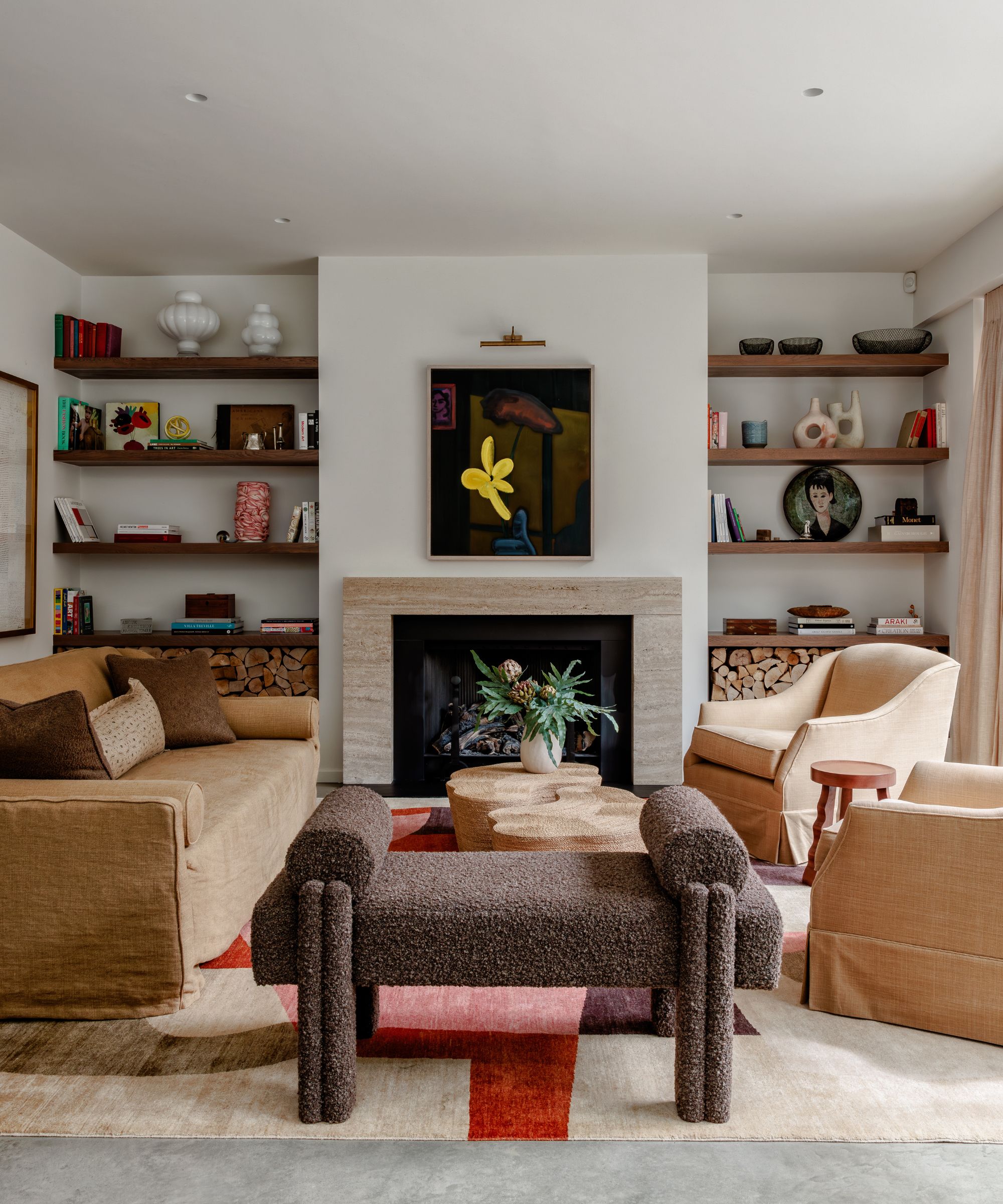Modern, neutral-toned living room with a fireplace, built-in wooden shelves filled with books and decor, a beige sofa, a dark textured bench, and an abstract yellow floral painting above the mantel.