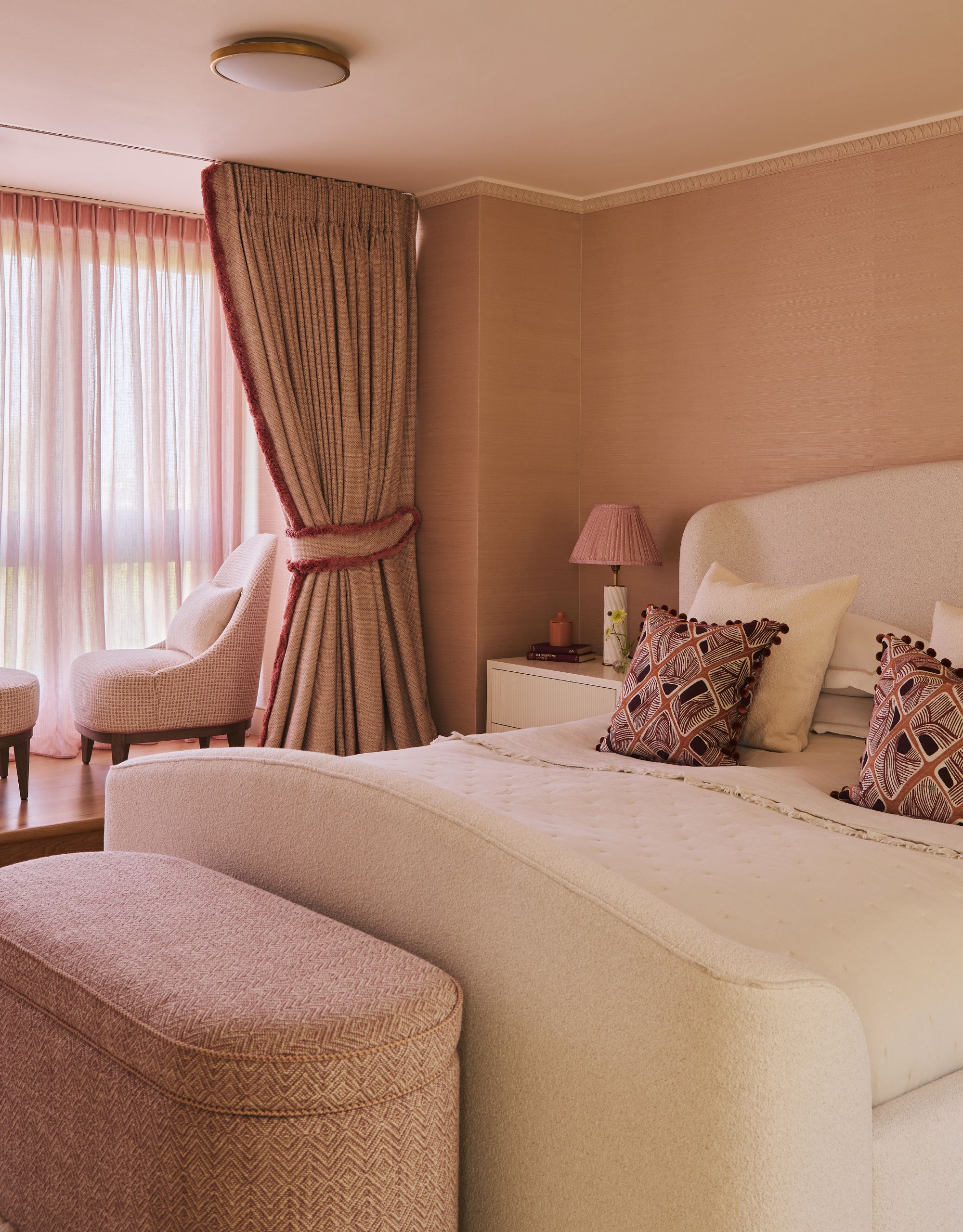 A bedroom with pink walls, curtains and storage bench, and a bed with a white bedframe and bedding and some patterned scatter cushions; there's two accent chairs behind the curtains by the window