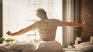 A man stretches out his arms in front of an open window after waking up early