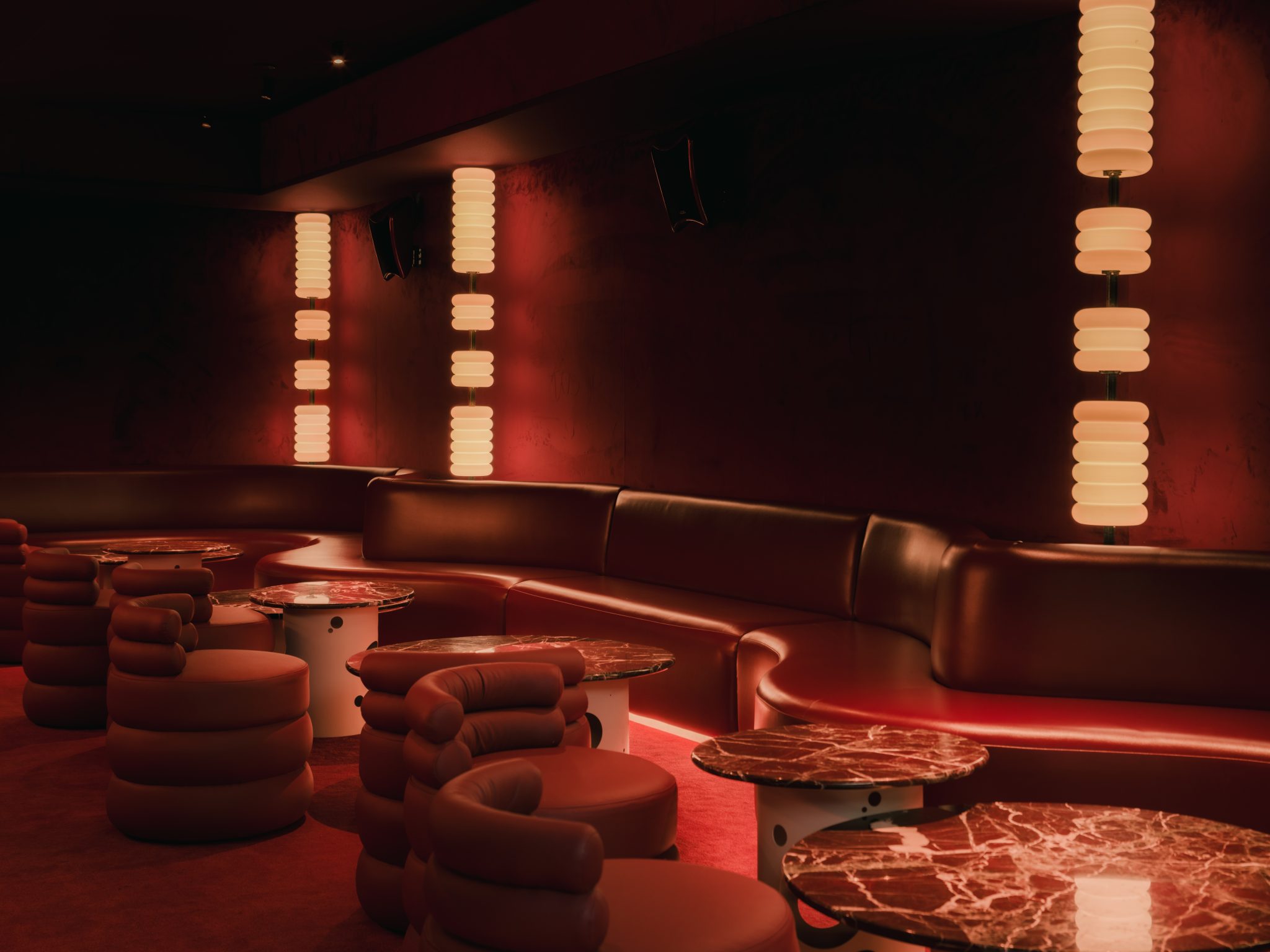 A dimly lit bar with red and orange-tinted interiors sculpted in wood, chrome, lacquer, velvet, and marble features sinuous banquettes, atmospheric sconces, geometrical skylights, and refined details.
