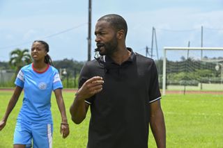 Emmanuel Warner (Bobby Gordon) is refereeing a woman's football match. He is dressed all in black and blowing his whistle. There is a player behind him in a blue kit with white stripes on the sleeves of her jersey and the sides of her shorts.