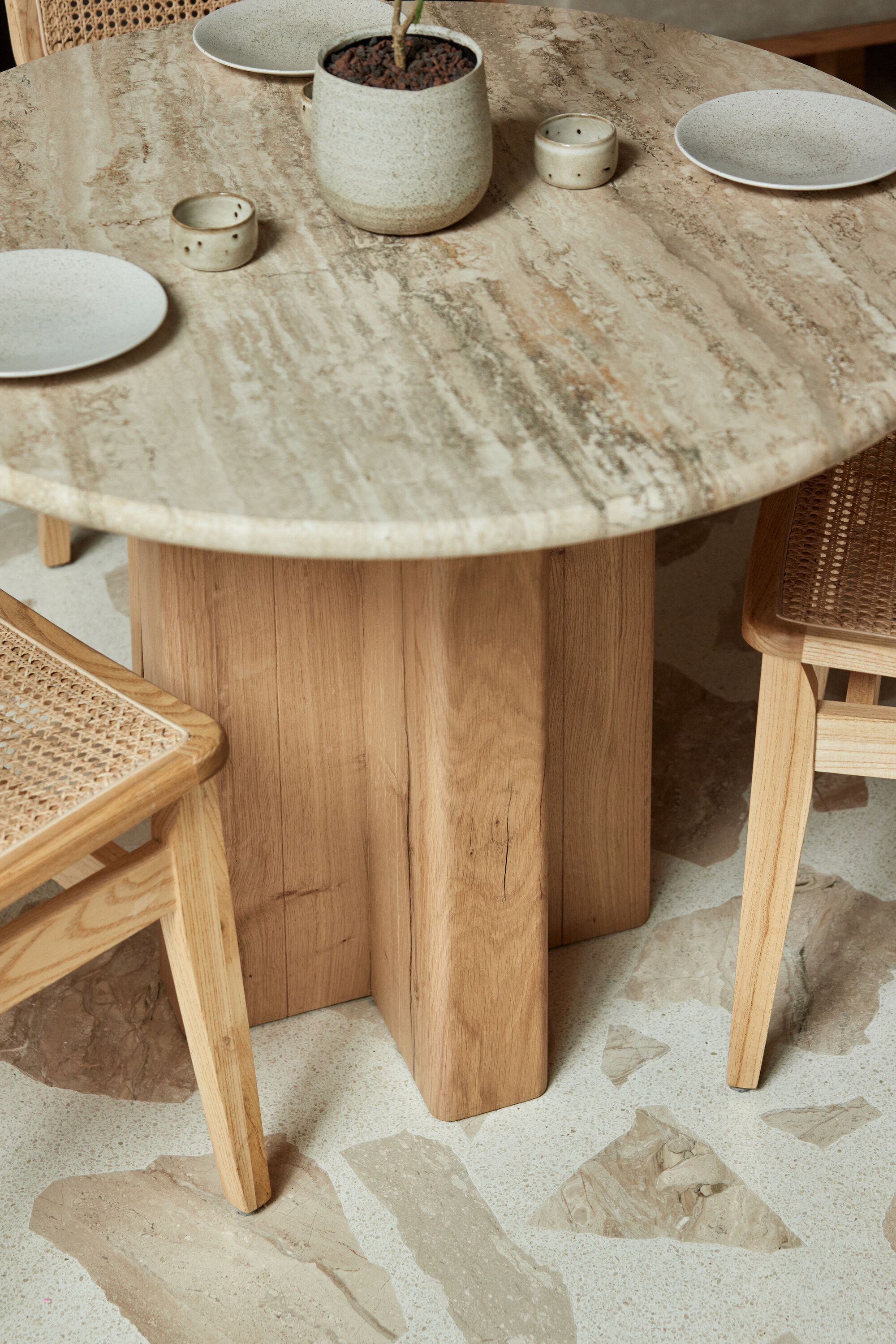 Close-up image of a round dining table with a marble top and a wooden base. There are three plates on the table, and wooden and rattan chairs are placed around the table. The floors are a blush pink and white palladiana terrazzo tile.