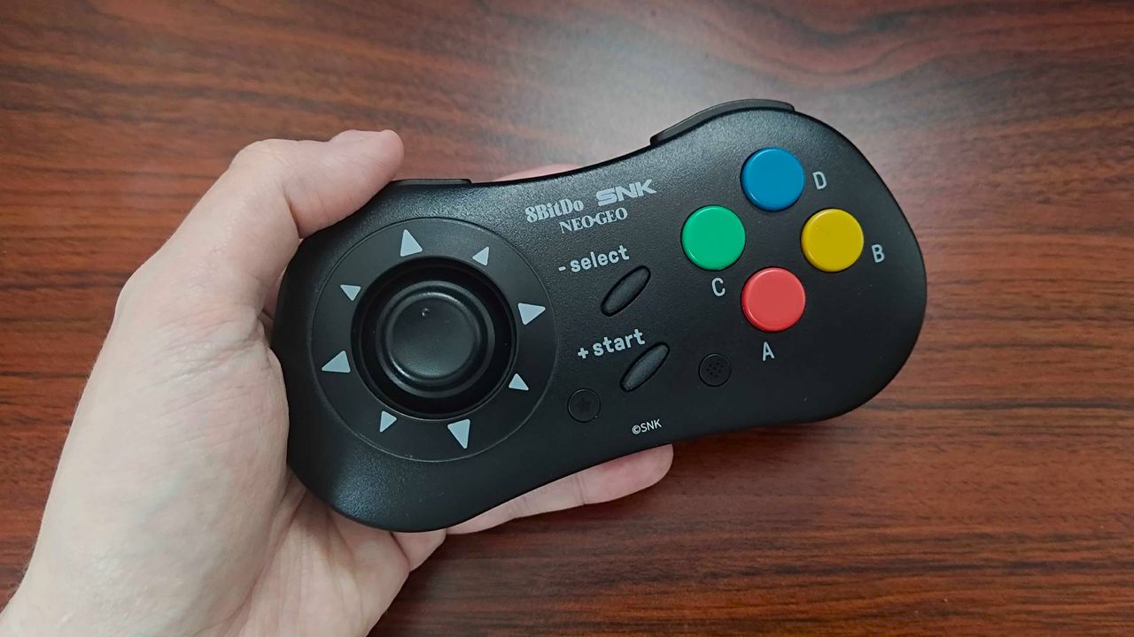 Hand holding 8bitdo Neo Geo controller with woodgrain desk.