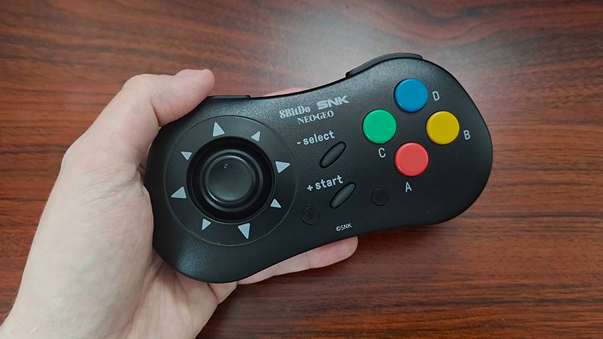 Hand holding 8bitdo Neo Geo controller with woodgrain desk.