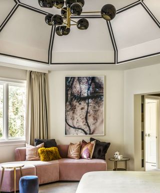 Large bedroom with unique, domed ceiling with black paint outline, cream painted walls and curtains, pink sofa, cream carpet, modern artwork on wall, cream bed