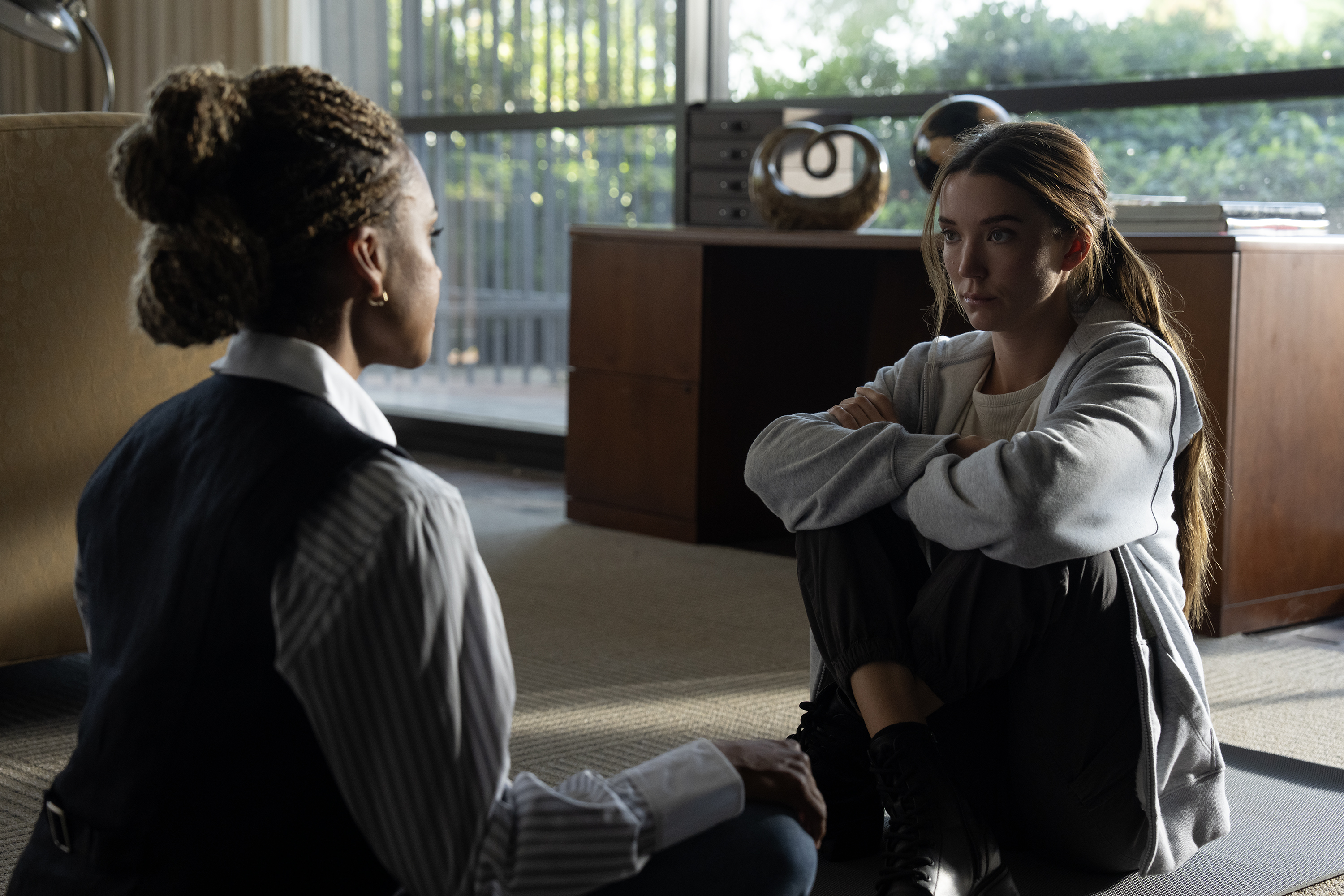 A black woman in a suit (Ryan Michelle Bathe as Stacy) sits on the floor of an office with a white woman in sweats (Nicole Brydon Bloom as Jane), in a still from 'Paradise' season 2.