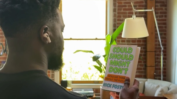 A man seen from behind and to the side, holding a up book while wearing an Apple AirPod earbud in one ear.