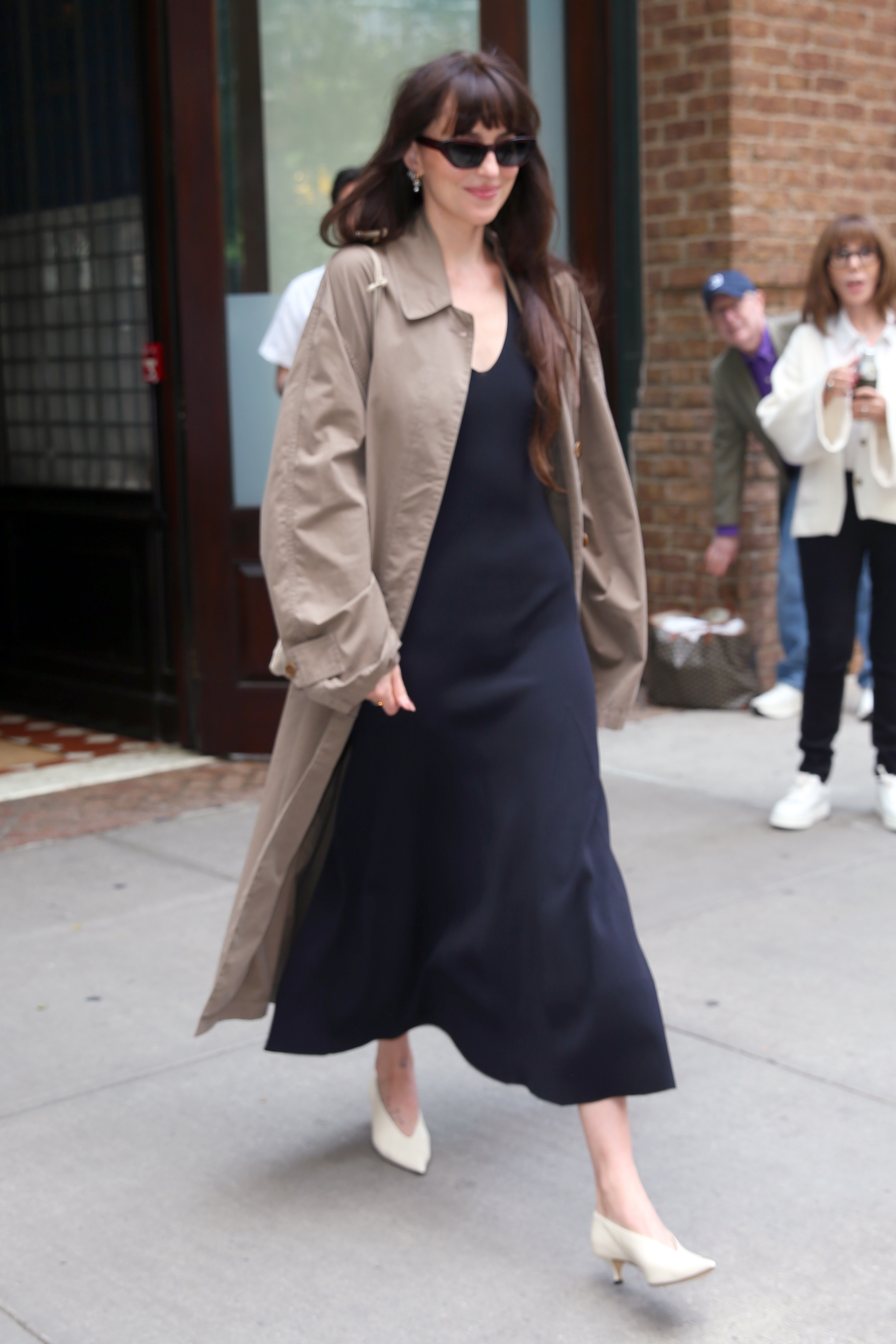 Dakota Johnson steps outside in New York wearing cream heels with a navy dress and a dark beige trench coat.