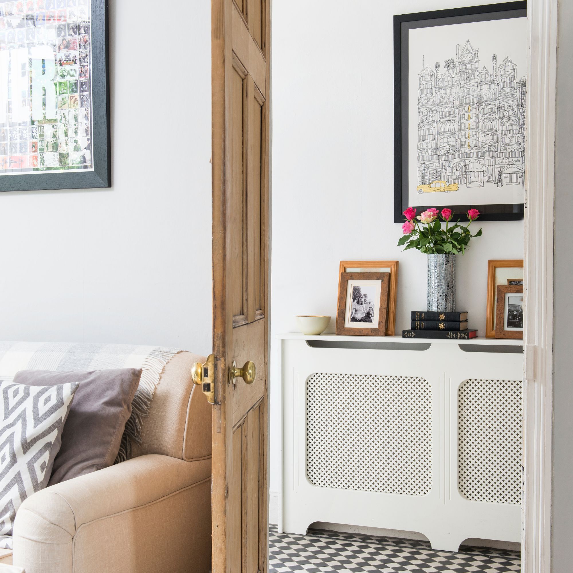 Doorway from living room to hallway, where you can see a cream radiator cover, with framed pictures, books and vase with flowers on top, a framed print on the wall and a black and white checkerboard floor