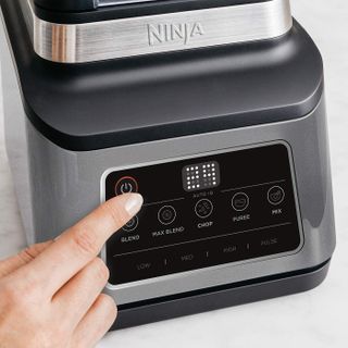 A woman pressing the operation buttons on a Ninja 3-in-1 Food Processor on a white kitchen counter
