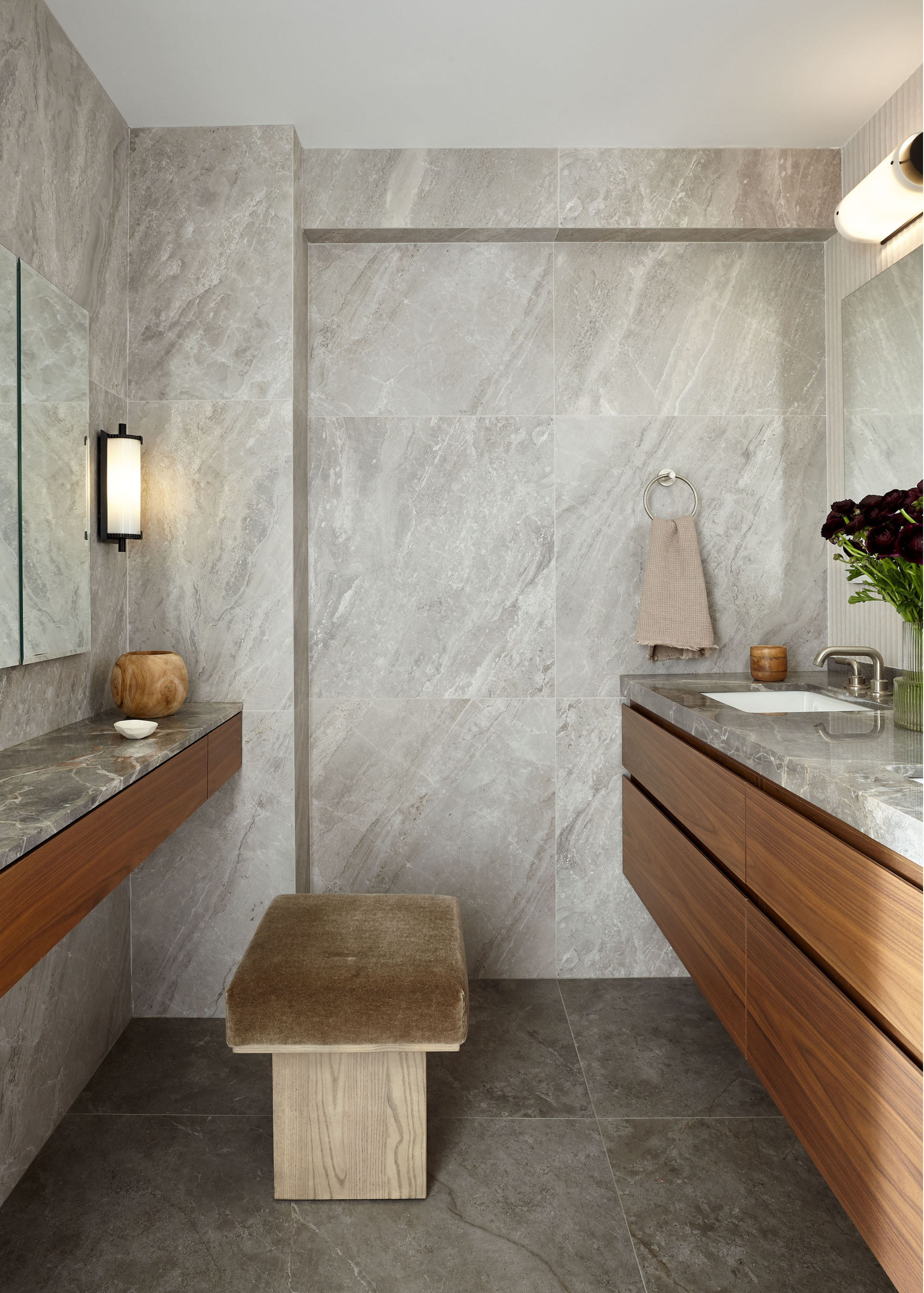 A grey tiled bathroom in a galley layout with wooden vanity storage beneath the grey marble top and a wood bench topped with olive velvet to sit on at one side