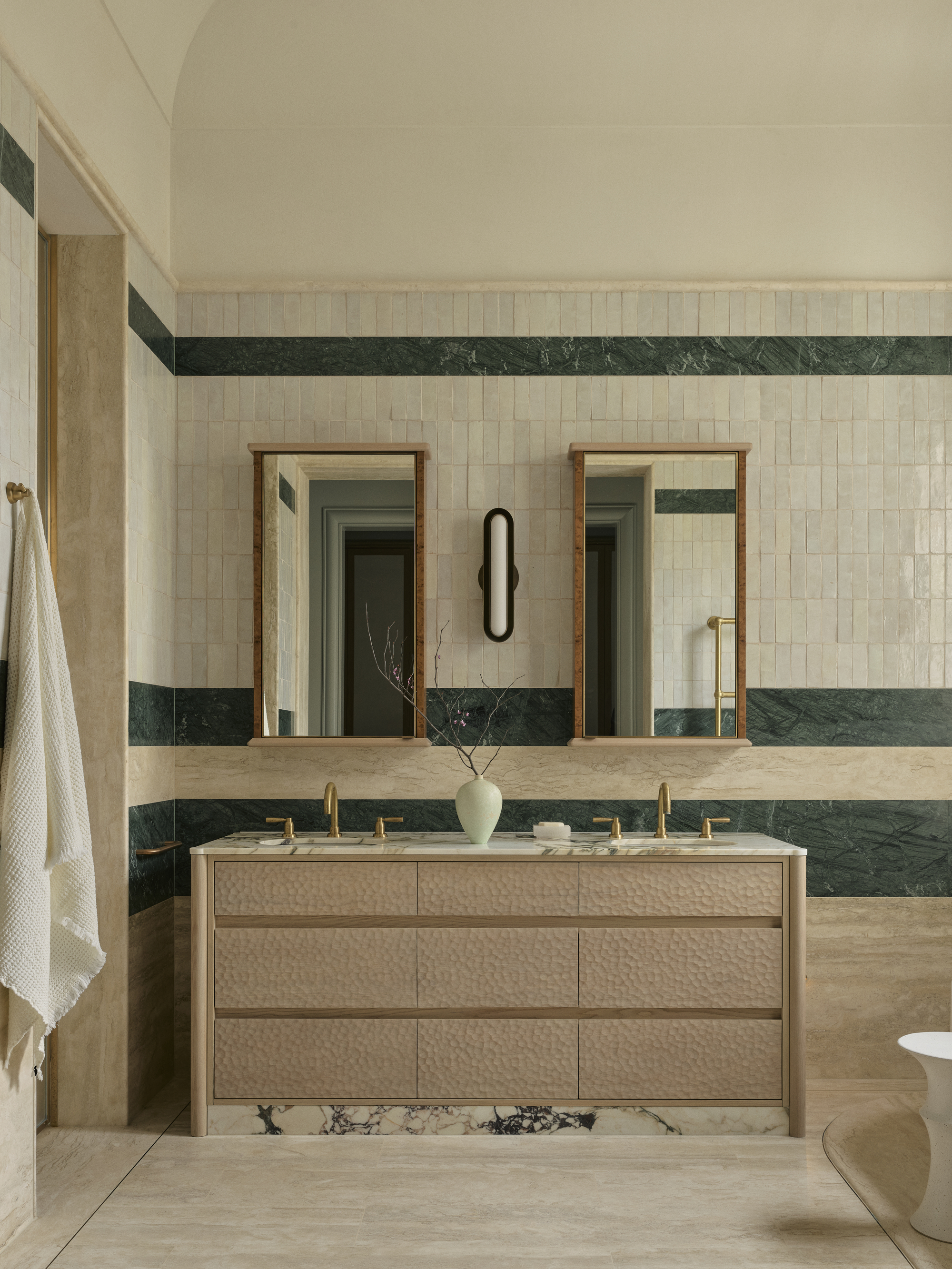 modern bathroom with travertine floors, green and white wall tiles, marble vanity with blonde carved timber vanity, two mirrors, wall sconce, brass taps, and a vase with a branch on the vanity