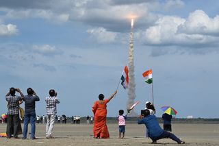 People watch a rocket launching in the distance and wave Indian flags.