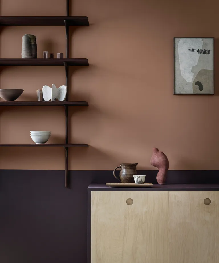 A two toned wall with light brown paint on the upper and dark brown paint on the bottom, a dark woork open shelving unit with small decor and a light wood side table.