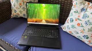 A black laptop rests on a blue cushion between floral pillows on a wicker sofa; its screen shows a sunlit path through a vivid green forest.