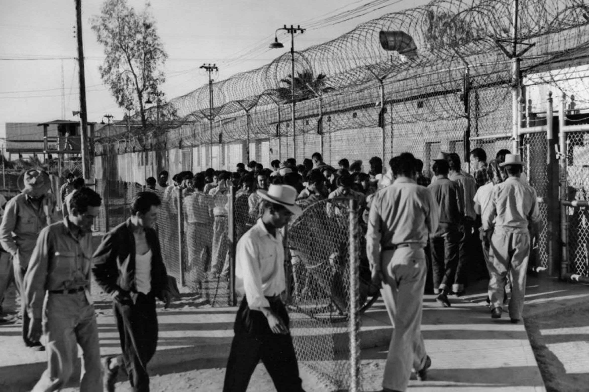 A large crowd of men stands behind a barbed-wire-topped chain-link fence at a detention compound, with more men walking in the foreground.