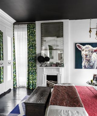 Contemporary bedroom with black painted ceiling, white painted walls, painted white fireplace, botanical curtains, black painted floor, bed with black and pink throw, lamb painting on the wall
