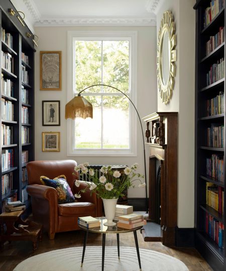 Reading nook with large windows, bookshelves on either side, a leather brown armchair, a vintage floor lamp with a fringed shade, and a round coffee table holding books and a vase of flowers.